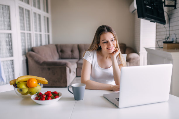 在家里的厨房里，一位微笑着端着咖啡的年轻女子正在用笔记本电脑进行视频聊天图片下载