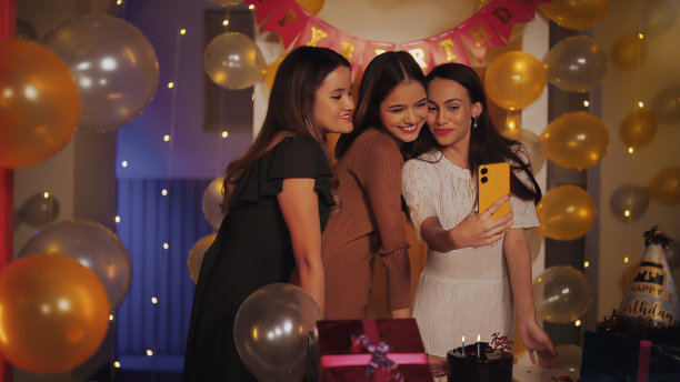 印度年轻漂亮的z世代女性朋友站着用手机拍滑稽的脸摆自拍照片享受室内装饰的家。现代青少年影响女孩拿着手机看cam制作内容视频拍摄庆祝生日的房子图片下载