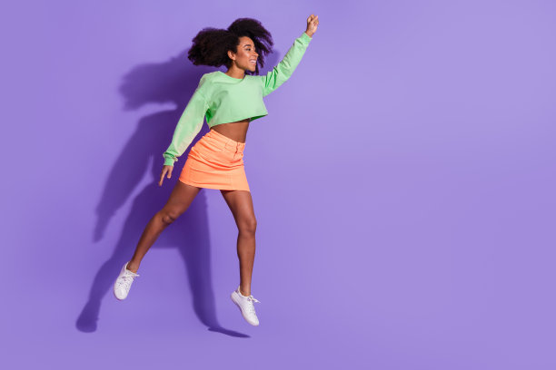 年轻有趣的跳跃女孩的全身照片，波浪状的头发在绿色的顶部，举起拳头，超级英雄孤立在紫色的背景图片下载