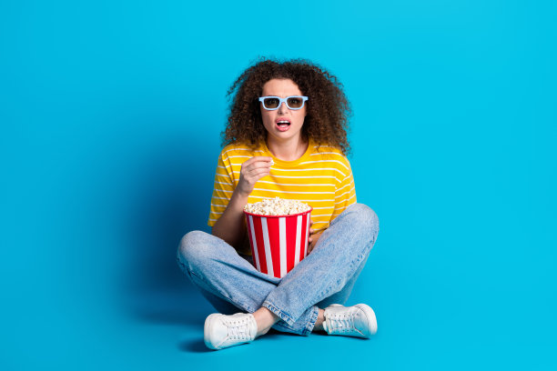 漂亮的年轻女孩的全尺寸照片坐在地板上看电影吃爆米花穿时髦的黄色条纹装孤立的蓝色背景图片下载