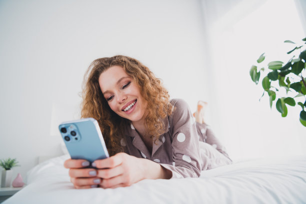 漂亮的年轻女孩的全尺寸照片躺在床上使用电话，穿睡衣明亮的室内卧室公寓室内图片下载