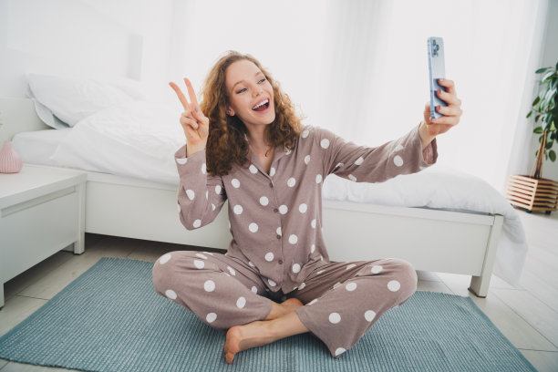 照片漂亮的年轻女孩做自拍手机v字手势穿着睡衣明亮的卧室公寓室内图片下载
