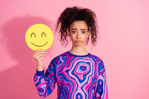 照片中压力大、情绪低落的女人穿着印花运动衫，手里拿着笑脸，空无一物，孤立的粉红色背景图片下载