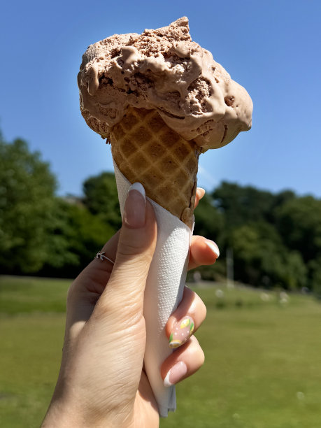 照片中，无法辨认的女性拿着用餐巾包裹的华夫饼筒，一勺融化的巧克力冰淇淋，涂着柠檬水果和柑橘片的指甲油，阳光明媚的公园背景，重点放在前景上图片下载