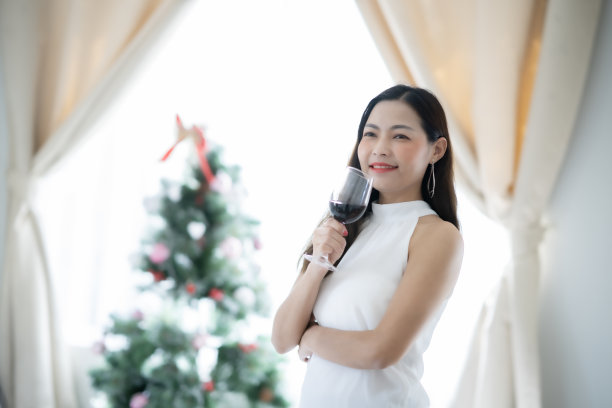 亚洲妇女端着一杯红酒站在客厅里，微笑着与圣诞树庆祝幸福。图片下载