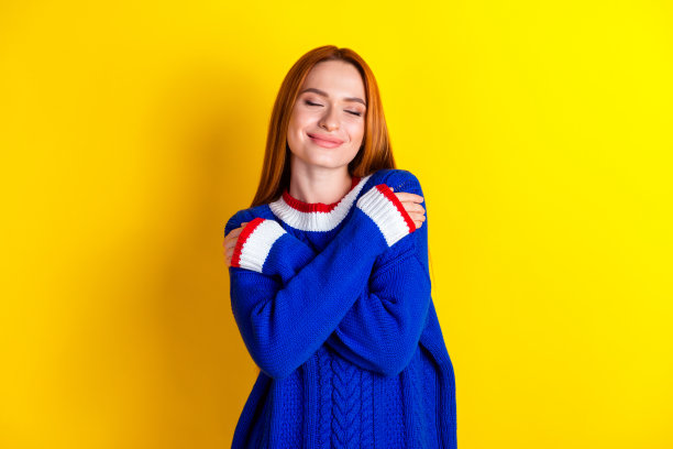 照片红头发的模特年轻开朗的女人在蓝色针织毛衣拥抱自己舒适的衣服孤立的黄色背景图片下载