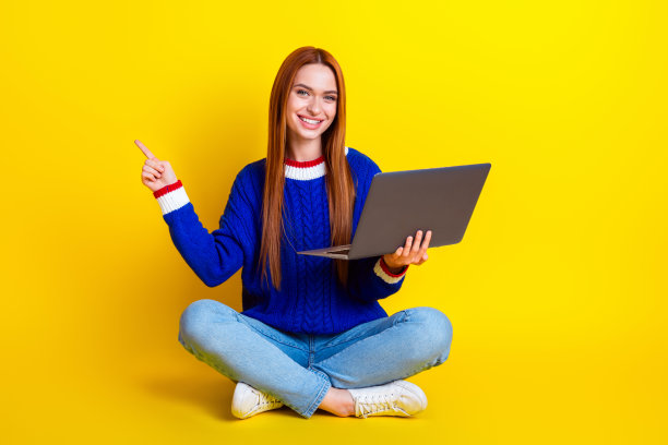 照片中的年轻快乐的女人穿着蓝色时尚的套头衫和牛仔裤，使用笔记本电脑工作，推荐有用的应用程序孤立的黄色背景图片下载