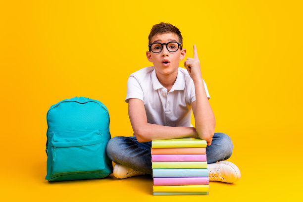 搞笑兴奋的男孩全身照片穿白色t恤书包看书指向空的空间孤立的黄色背景图片下载