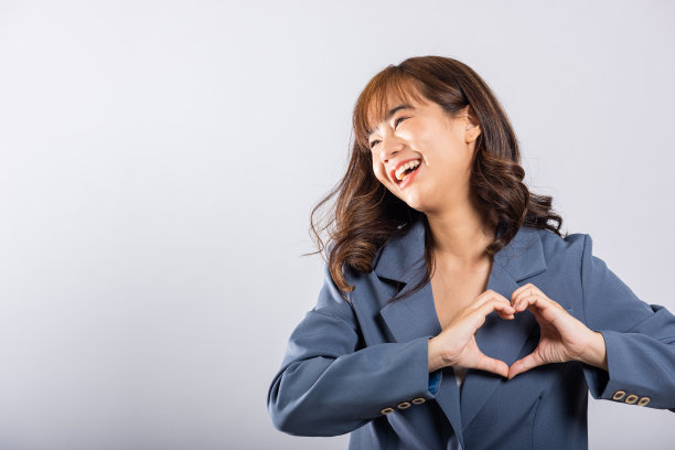 一位自信的商务女性穿着西装，微笑着在白色的背景上画出两只手的心形图案。一个美丽的年轻女性表达爱情的亚洲肖像图片下载