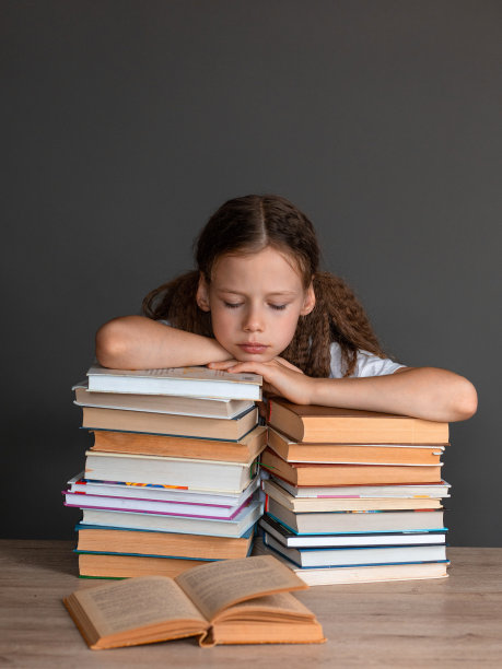 一个穿着白色t恤的女孩坐在一堆书上，躺在灰色的背景上看书。学习和作业。孩子在学校的压力。准备考试。学校科目导师。学校里信息过多。额外的教育。图片下载