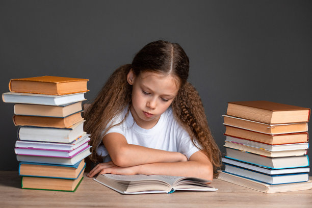 一个穿着白色t恤的女孩坐在桌子旁，在灰色的背景上看书。学习和作业。孩子在学校的压力。准备考试。学校科目导师。学校里信息过多。额外的教育。图片下载