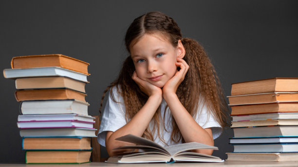 一个穿着白色t恤的女孩坐在书中间的桌子旁读书，用手枕着头，枕在灰色的背景上。学习和作业。孩子在学校的压力。准备考试。学校科目导师。学校里信息过多。图片下载