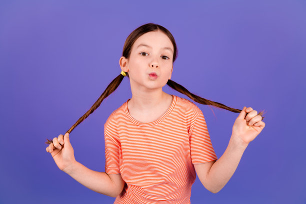 照片肖像可爱的小女孩拉头发尾巴亲吻嘴唇穿着时尚的红色服装孤立的紫罗兰色背景图片下载