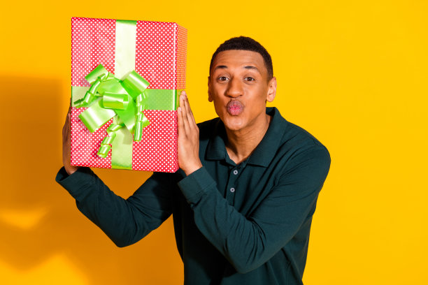 漂亮的年轻男性的照片肖像拿着礼盒亲吻嘴唇穿着时髦的绿色服装孤立的黄色背景图片下载