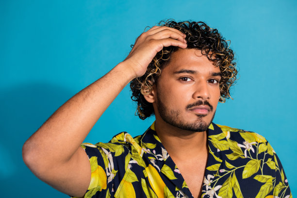 照片英俊的都市美男，留着胡子，波浪发型，穿着印花衬衫，手放在头上，孤立在蓝色背景上图片下载