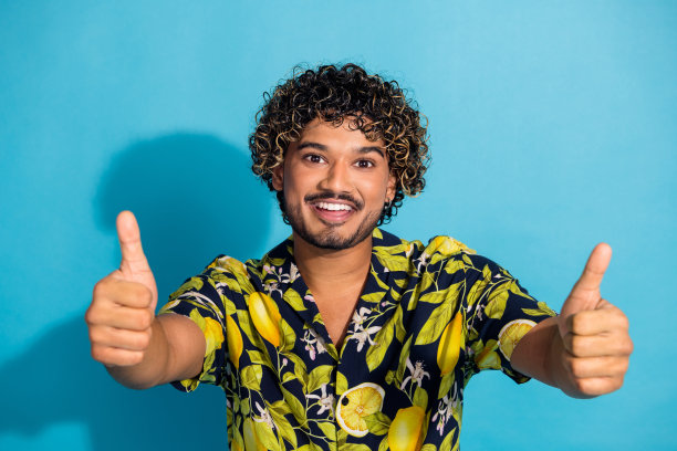 照片满意乐观的家伙与胡子波浪发型穿着印花衬衫显示你的大拇指孤立的蓝色背景图片下载