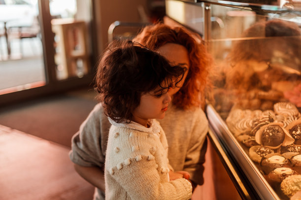 可爱的小卷发女孩3岁与一个年轻的红头发的妈妈在面包店的柜台上选择糕点。咖啡馆。图片下载