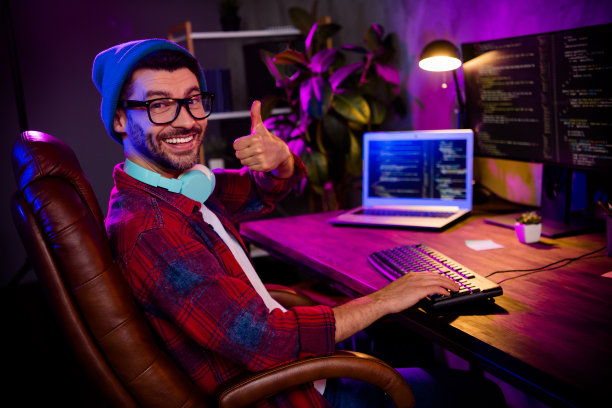 照片自信微笑的程序员戴着帽子，戴着眼镜，大拇指朝上，在工作场所的工作站阁楼里工作图片下载