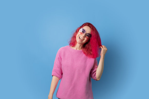 在蓝色背景下，戴着太阳镜的粉色头发的女人，穿着时髦的运动衫，时髦的表情。图片下载