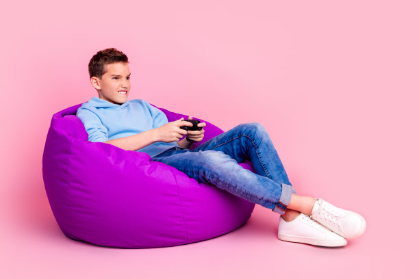 有趣的十几岁男孩的全尺寸照片豆袋playstation玩视频游戏兴奋穿着时髦的蓝色服装孤立在粉红色的背景图片下载