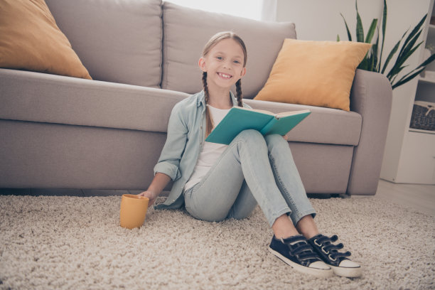 可爱小女孩的全尺寸照片坐在地毯地板穿衬衫学习看书喝茶现代公寓室内图片下载