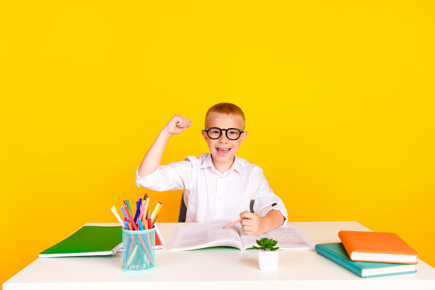 照片中聪明开朗兴奋的男孩穿着白衬衫坐在书桌上做作业，孤立在黄色背景上图片下载
