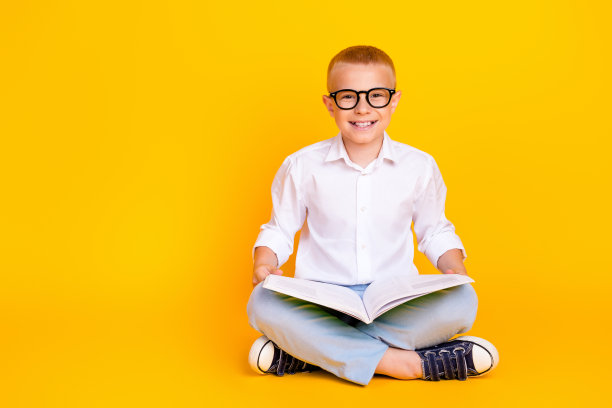照片中开朗聪明的男孩穿着白衬衫，坐在那里看书，黄色的背景空无一物图片下载