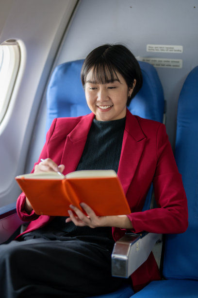 一个微笑的女人坐在飞机窗口，全神贯注地在她的橙色笔记本上写着笔记。图片下载