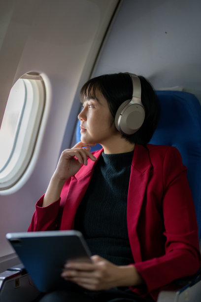 一位穿着红色夹克，头戴耳机的深思熟虑的女士手持平板电脑向飞机窗外望去。图片下载