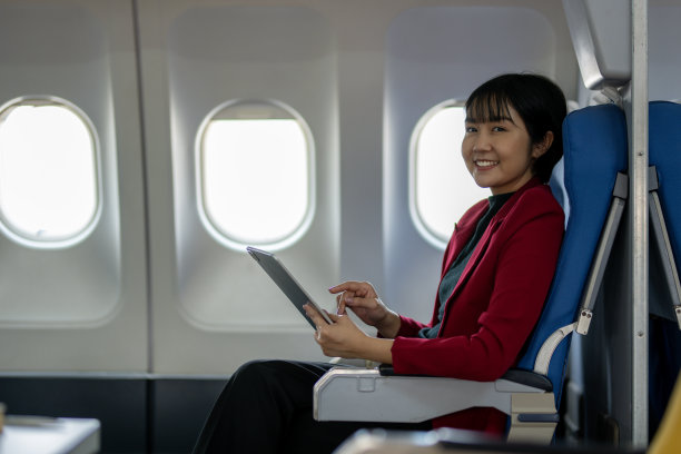 穿着黄色西装、微笑着的女商人在飞机上高效地工作着，桌上放着笔记本电脑。图片下载