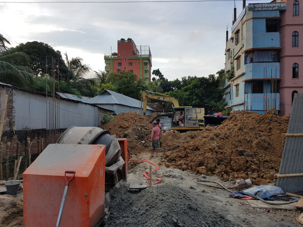 2024年8月5日，孟加拉国达卡。这张照片显示了一个建筑工地，有一大堆土，一辆黄色的挖掘机，一辆红色的自卸卡车，以及一些穿着蓝色衬衫的工人。图片下载