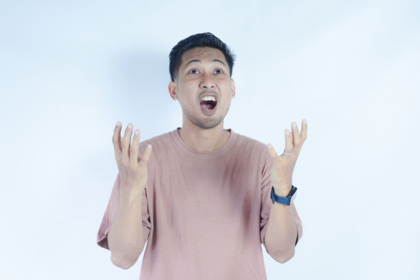 一名身穿粉色t恤的亚洲男子，站在朴素的背景下，脸上带着惊讶的表情，双手举到脸附近。这张照片捕捉到了震惊或惊讶的瞬间。查找图片下载