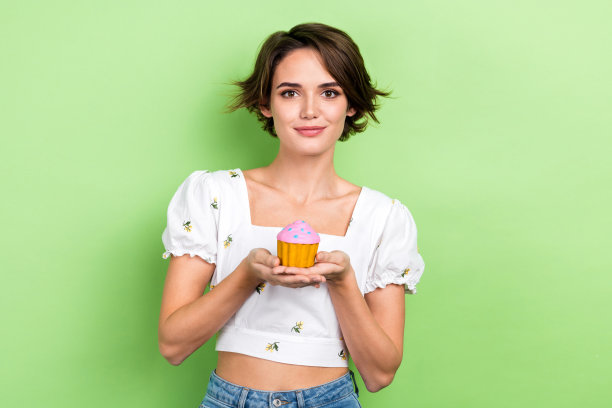 照片中时髦漂亮的女士穿着白色上衣，双臂抱着小纸杯蛋糕，孤立的绿色背景图片下载