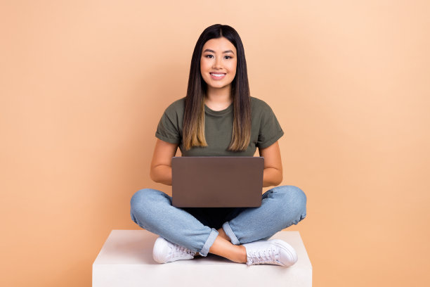 迷人的中国女人的全尺寸身体照片，黑发坐在平台上，苹果macbook it技术孤立在米色背景上图片下载