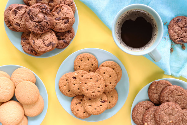 一盘刚烤好的自制混合饼干的特写图片，周围满是黑咖啡，蓝色马克杯，双层巧克力饼干，巧克力饼干，全麦饼干和酥饼圆，黄色背景，高架视图图片下载