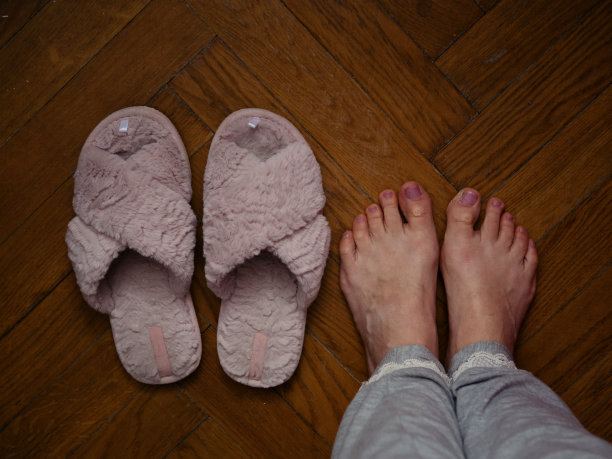 正上方的水平特写，一个穿着灰色睡衣的女人赤脚站在镶木地板上，旁边是粉红色的绒毛拖鞋，脚部有瑕疵。偷拍的家庭照片。图片下载