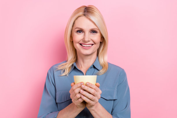 迷人的高级商务女士的照片拿着热杯咖啡休息时间穿着聪明休闲的蓝色衬衫孤立的粉红色背景图片下载