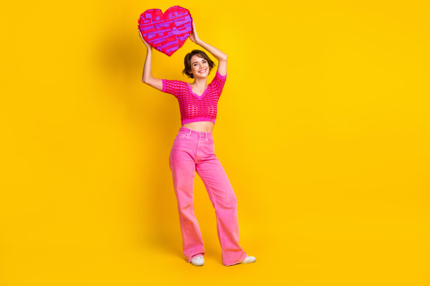 愉快可爱的女人穿着粉红色针织裤的全身照片，手臂在黄色背景上抱着大心脏图片下载