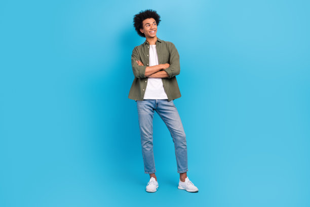 年轻模特的全身照片，帅哥交叉双手，自信地穿着卡其色衬衫牛仔裤，在蓝色背景下看起来很孤立图片下载
