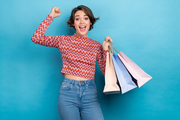 照片肖像的漂亮的年轻女孩举起拳头购物袋巨大的销售穿着时尚的条纹服装孤立的蓝色背景图片下载