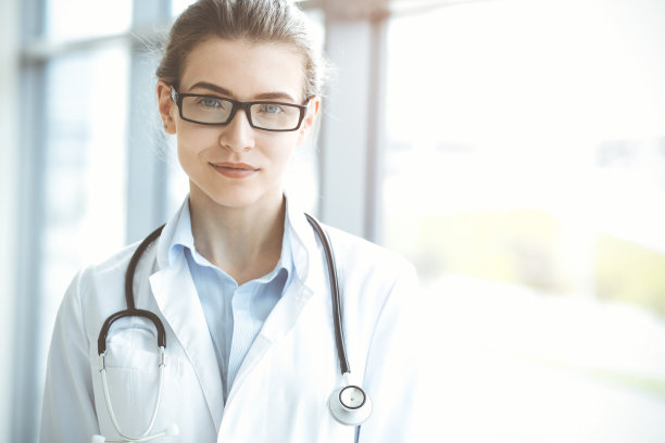 在阳光明媚的医院里，带着听诊器的美女医生微笑着，随时准备帮助病人。医学的概念图片下载