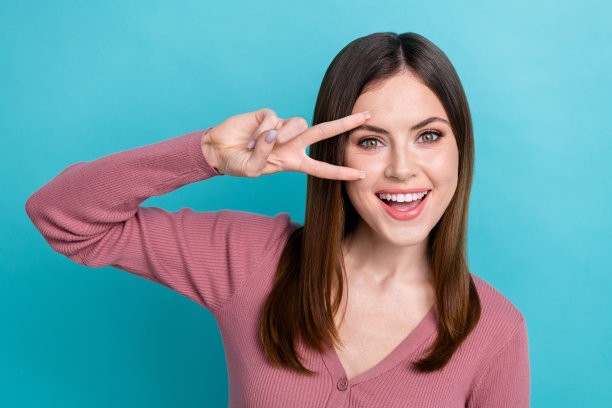 照片中的年轻女孩穿着粉红色的衬衫，遮住脸v字符号手指你好近眼睛好莱坞的牙齿微笑孤立的青色背景图片下载