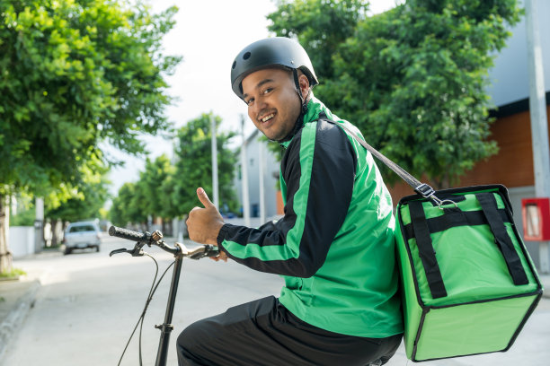 肖像骑手送餐员穿着绿色制服戴着头盔骑着自行车送餐给顾客。快乐快递员用绿色背包将货物运送给客户。图片下载