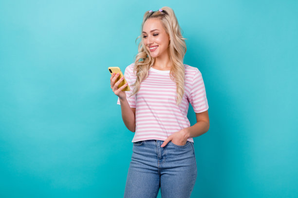 照片微笑满意的博主年轻女孩金发卷发阅读手机选项界面更新孤立的海蓝宝石色背景图片下载
