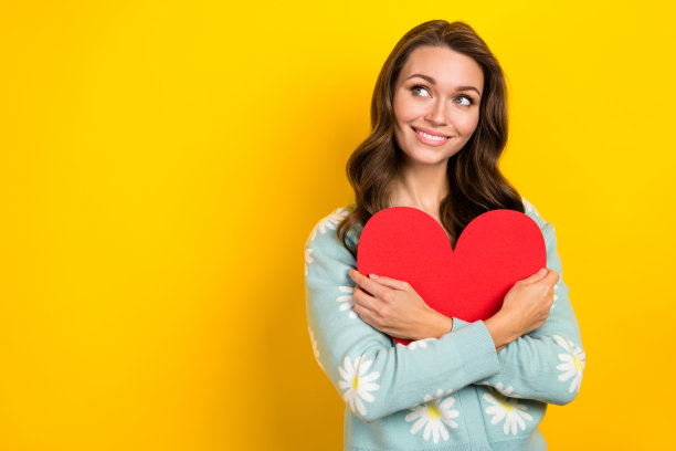 照片的梦想可爱的年轻女子青色套头衫雏菊花印刷拥抱红纸爱的心看空空间孤立在黄色的背景图片下载