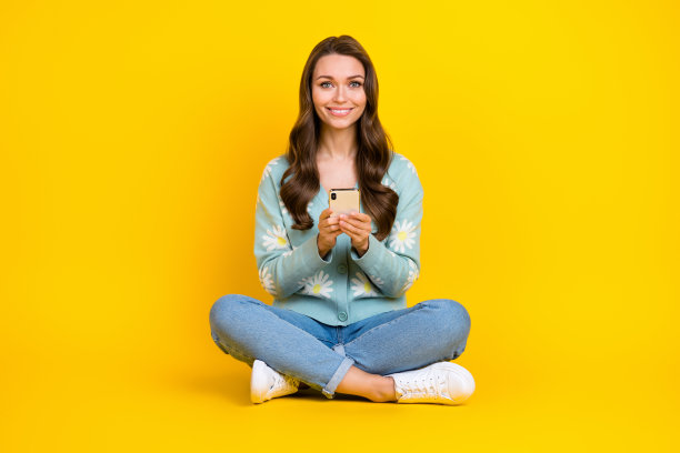 年轻开朗迷人女孩的全身照片，穿着时髦的衣服，用智能手机预订黄色背景上孤立的新酒店图片下载