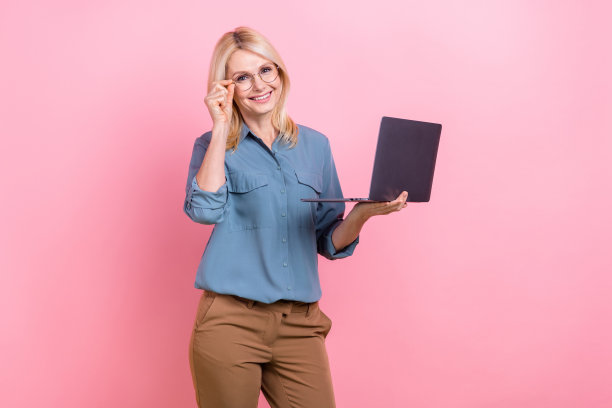 照片中微笑开朗的商务女士穿着蓝色衬衫棕色裤子拿着上网本触摸眼镜董事公司孤立在粉红色的背景图片下载