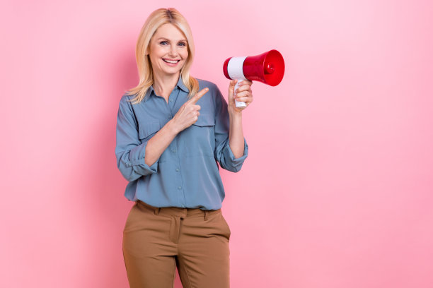照片中美丽的退休工人女士手持扬声器办公设备，穿着时髦休闲的蓝色衬衫孤立的粉红色背景图片下载