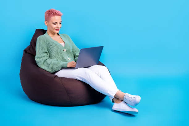 年轻的放松女孩的全身照片粉红色的头发坐在豆袋共享区域上网本编码java网页孤立的蓝色背景图片下载