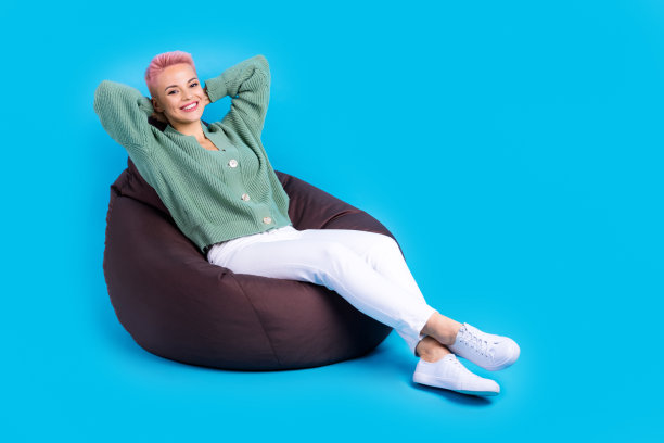 身材丰满的放松学生女生干部染了头发，穿着绿色的开襟羊毛衫躺着舒适的软豆袋度假孤立在蓝色的背景上图片下载